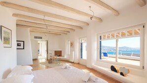 A spacious bedroom with a large bed, wooden beams on the ceiling, a view of the sea through a large window, and a balcony with seating. - Spacious Luxury Villa Delion-stunning sunset&bay view, infinity pool, by beaches (PAROS)