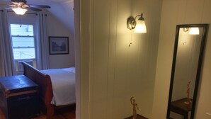 Een hotelkamer met een houten vloer, een bed met witte lakens, een tafeltje, een plafondventilator, een wandlamp en een spiegel.