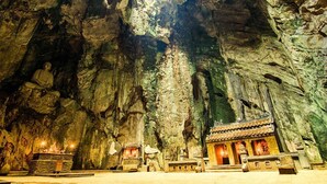 Un templo rupestre con una estatua de Buda sentado, un pequeño santuario y una construcción tradicional con techo de tejas.