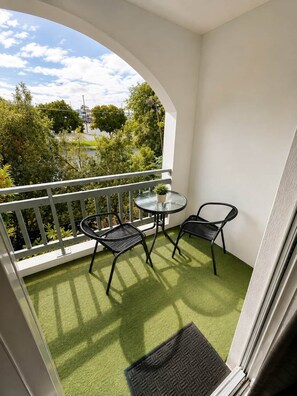 Un balcon avec une table en verre et deux chaises, une petite plante en pot et une vue sur les arbres et les bâtiments.