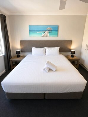 A hotel room with a large bed, two bedside lamps, a painting of a pier, and a rolled towel on the bed.