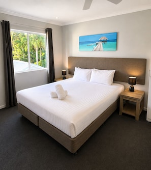 Een hotelkamer met een groot bed, een nachtkastje met een lamp, een schilderij van een strandlandschap en een raam met gordijnen.