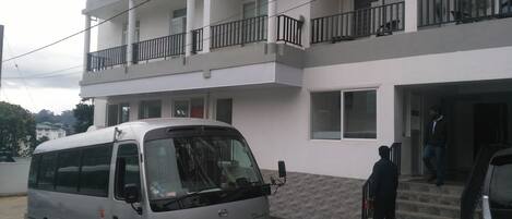 Un edificio blanco de varios pisos con balcones, una furgoneta estacionada y una persona de pie en la entrada.