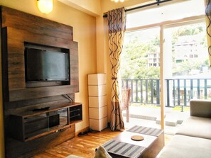 Un salon doté d’un téléviseur à écran plat fixé sur un mur en bois, d’un meuble blanc et d’un balcon offrant une vue sur la verdure et les bâtiments.
