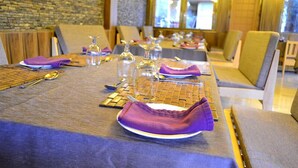 A dining area with tables set for multiple guests, featuring a stone wall and a view of a building through the window. - Hotel Migmar (Thimphu)