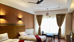 A hotel room with two beds, a desk, a chair, a ceiling fan, and a large window with curtains. - Hotel Migmar (Thimphu)