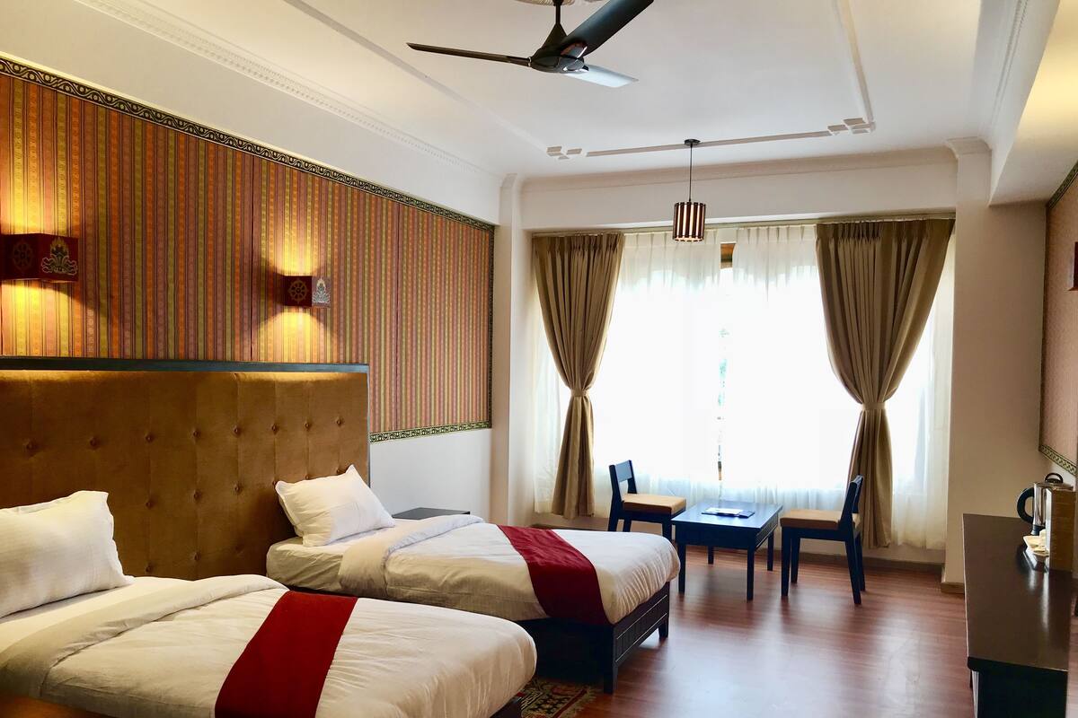 A hotel room with two beds, a desk, a chair, a ceiling fan, and a large window with curtains.