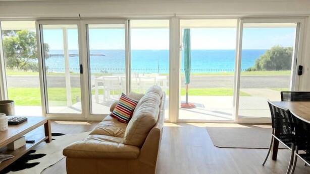A living room with a beige sofa, a small table, and a view of the ocean through large sliding glass doors. - Millpond - Penneshaw Beach (Penneshaw)