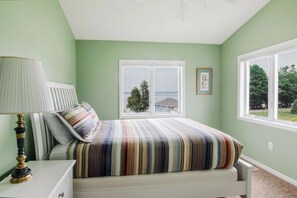 Quarto com cama, mesa de cabeceira com abajur, janela com vista para o exterior e quadro na parede.