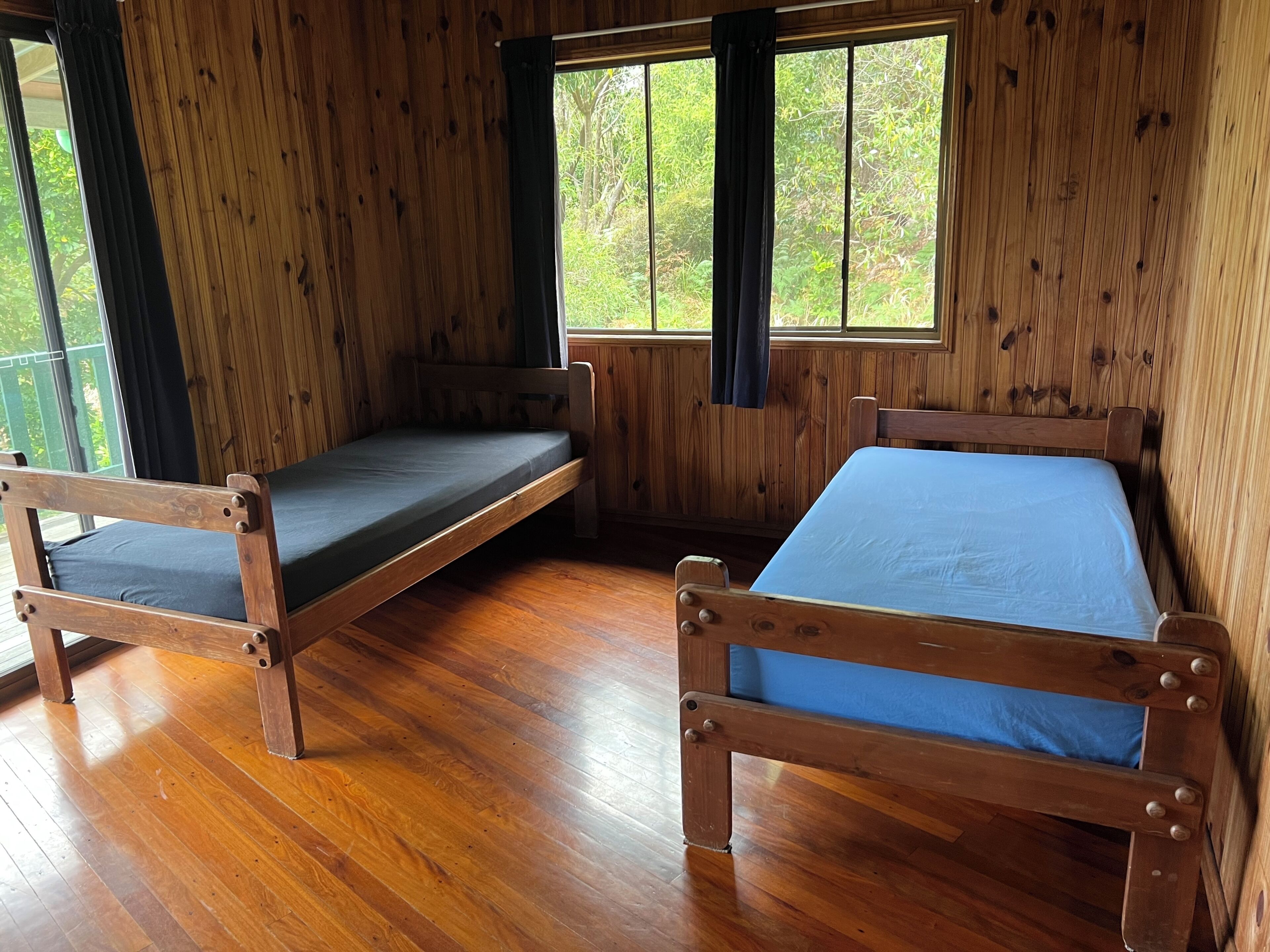 A cabin with two wooden beds, a wooden floor, and large windows with curtains.