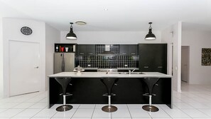 A modern kitchen with a central island, black bar stools, a stainless steel refrigerator, and a black backsplash. - Beautiful Gold Coast home, the perfect base for your Commonwealth Games stay (Mudgeeraba)