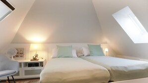Um quarto com teto inclinado, uma cama com duas almofadas, uma mesinha de cabeceira com abajur e uma cadeira.
