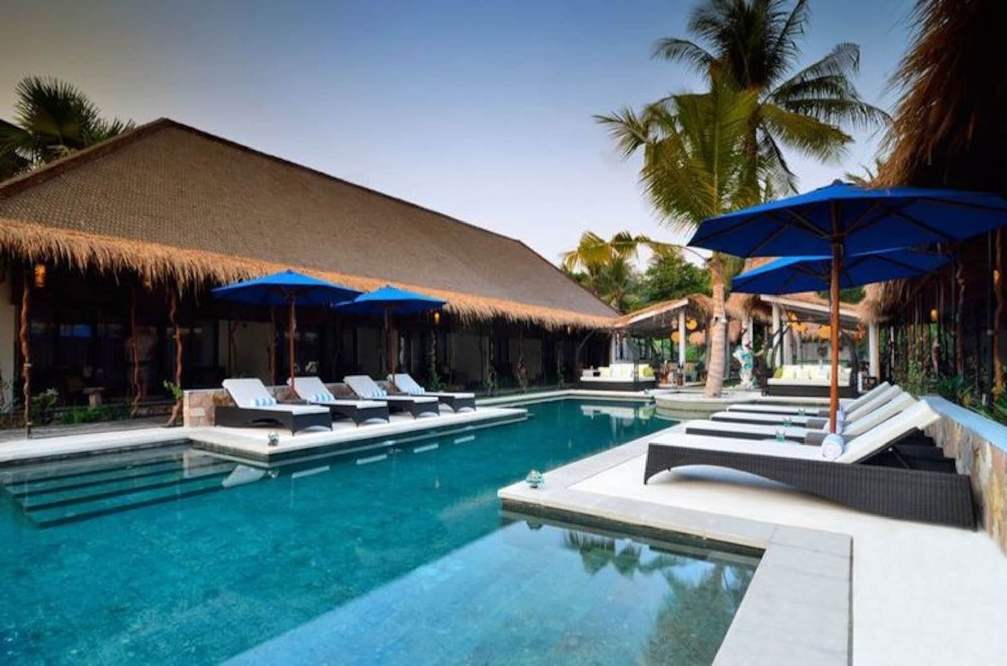 A pool area with lounge chairs and umbrellas, surrounded by palm trees and a thatched-roof structure.