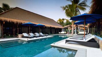 A pool area with lounge chairs and umbrellas, surrounded by palm trees and a thatched-roof structure.