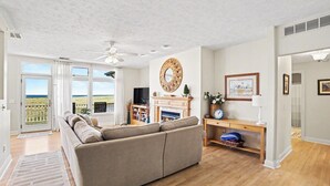 A living room with a sofa, TV, fireplace, and a view through large windows. - Second Floor Lake Michigan Condo with Sunset Views (Manistee)
