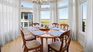 A dining area with a round table, six wooden chairs, a chandelier, and large windows with a view of the beach. - Second Floor Lake Michigan Condo with Sunset Views (Manistee)