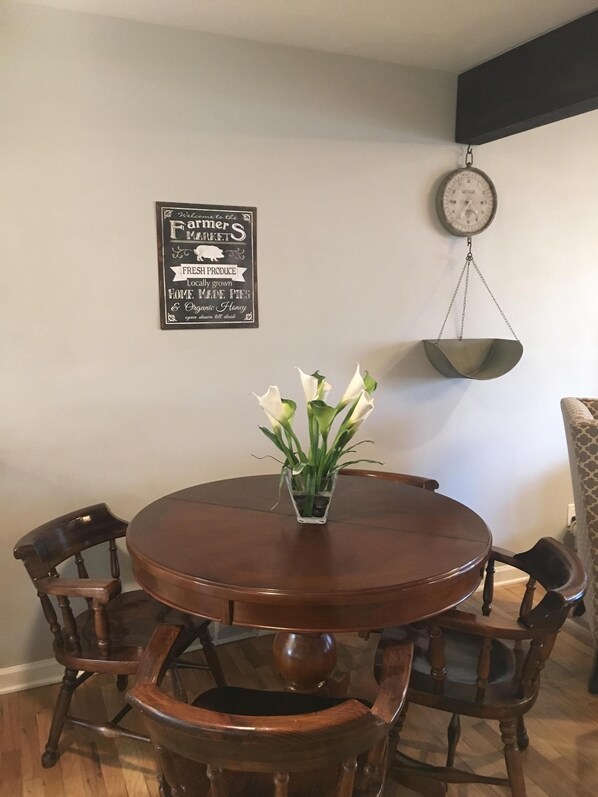 Un comedor con una mesa redonda de madera, cuatro sillas, un jarrón con flores blancas, un reloj de pared y un cartel sobre productos locales.