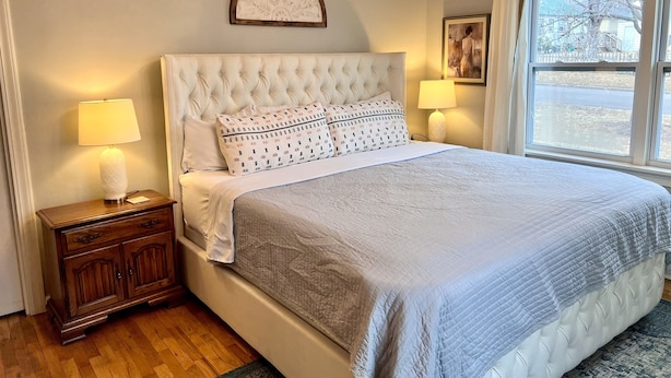 Un dormitorio con cabecera capitonada, una cama grande con almohadas estampadas, una mesita de noche con lámpara y una ventana con vistas al exterior.