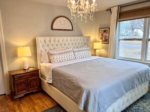 Un dormitorio con cabecera capitonada, una cama grande con almohadas estampadas, una mesita de noche con lámpara y una ventana con vistas al exterior.