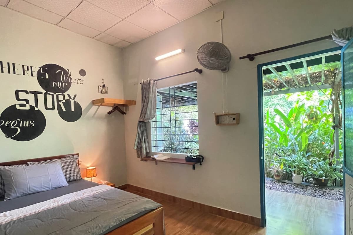 A bedroom with a bed, a wall-mounted fan, a window with curtains, and a view of greenery outside.