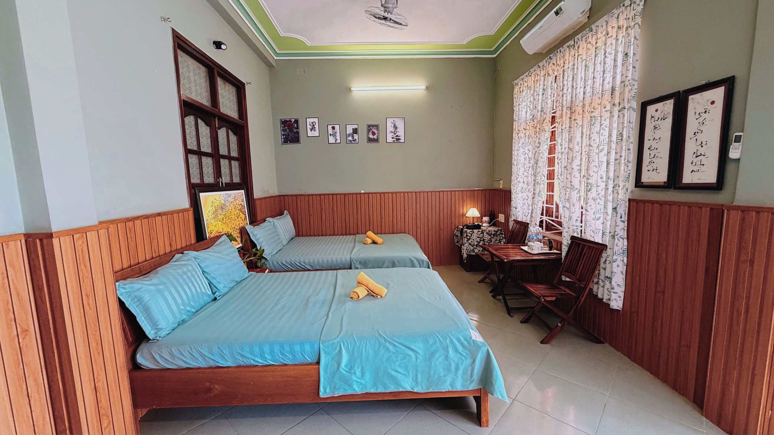 A room with two beds, a table with chairs, a lamp, and framed pictures on the wall.