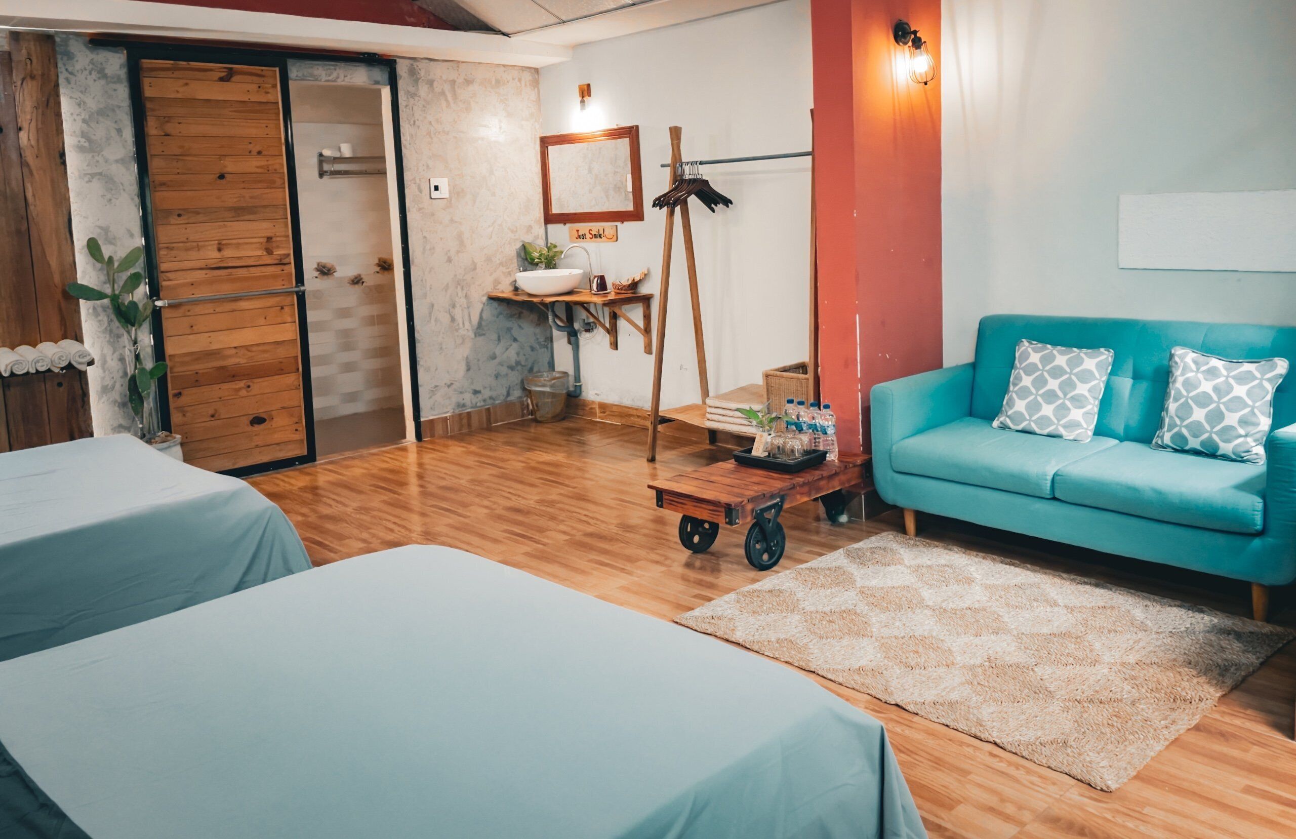 A room with wooden flooring, a teal sofa with patterned pillows, a wooden cart, a sink, and a mirror.