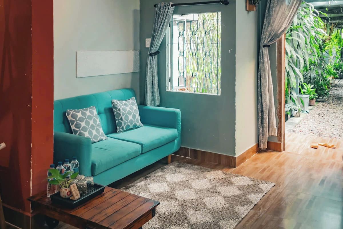 A teal sofa with patterned cushions, a wooden coffee table, a patterned rug, a window with metal bars, and a view of greenery outside.