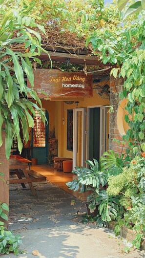 A rustic homestay with a wooden signboard, a covered entrance, and a garden area with various plants.