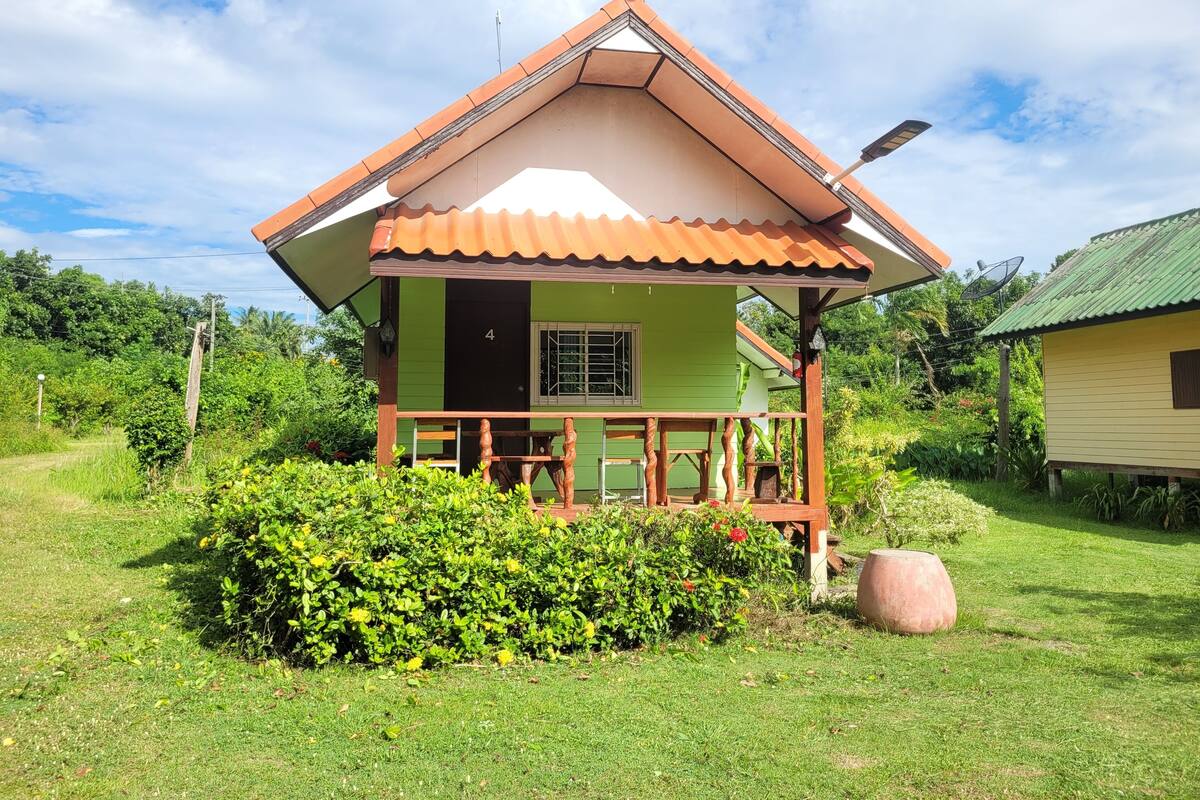 A small, single-story house with a green exterior and a brown roof. It has a porch with wooden railings and a satellite dish on the roof.
