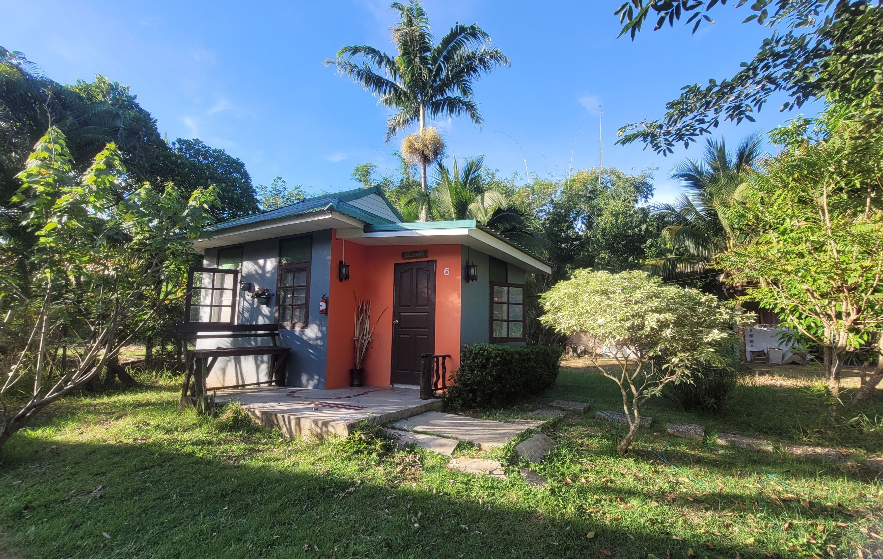 A small, single-story house with a red exterior and green roof, surrounded by lush greenery and palm trees.