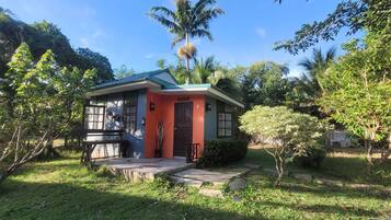 A small, single-story house with a red exterior and green roof, surrounded by lush greenery and palm trees.