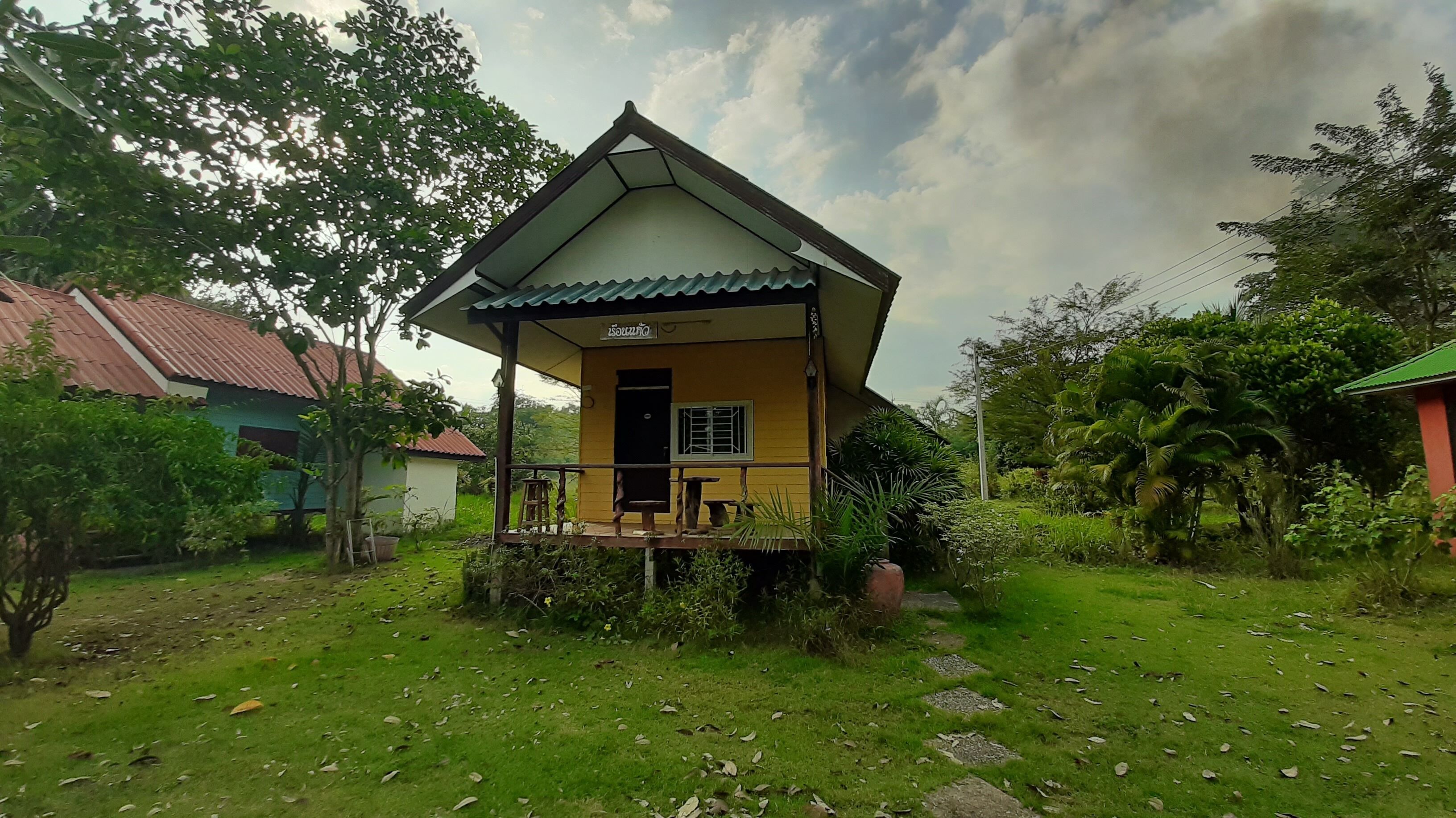 A small yellow house with a porch, surrounded by greenery and a red-roofed building in the background.