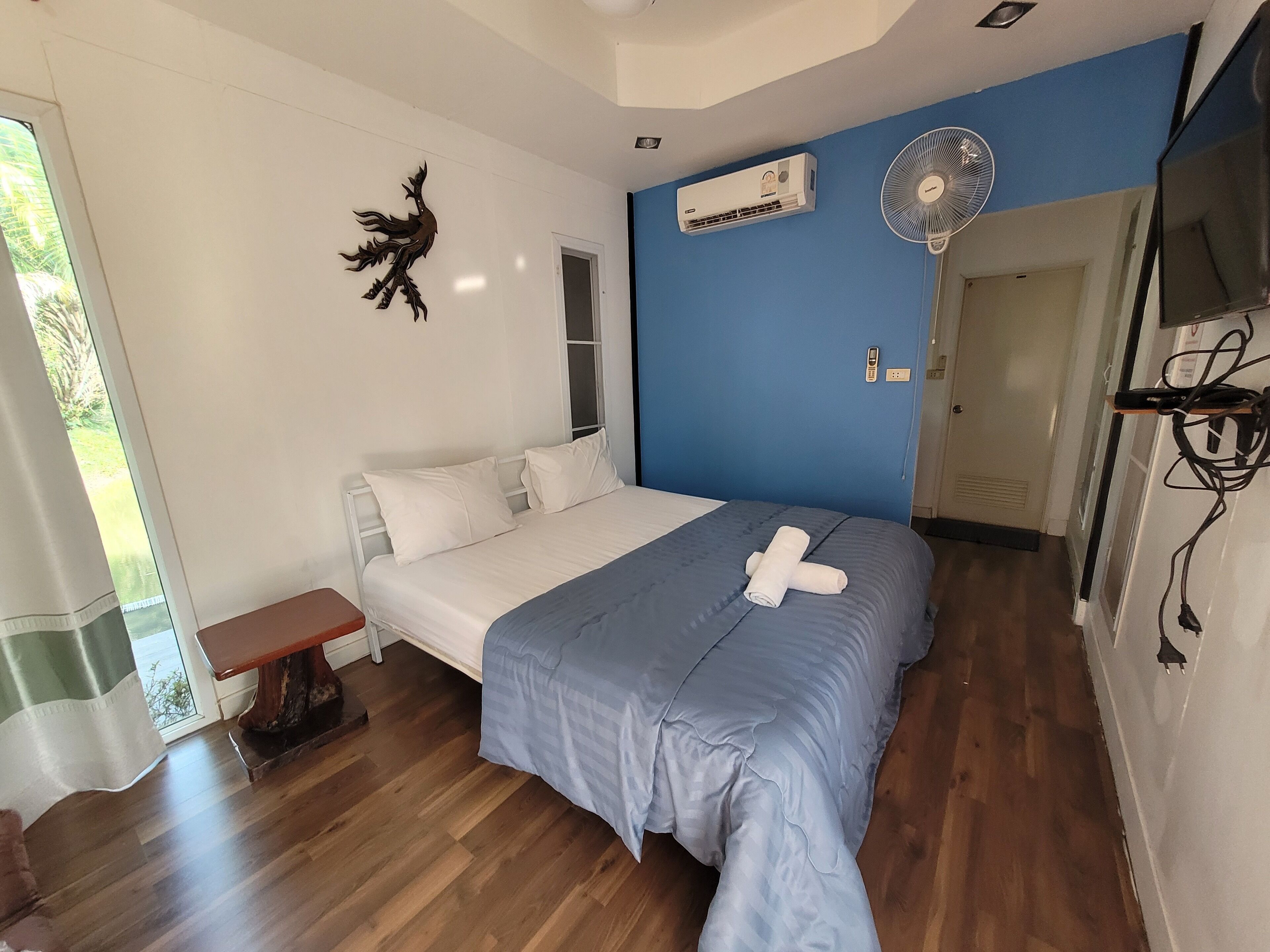 A bedroom with a bed, a wooden floor, a wall-mounted air conditioner, a ceiling fan, and a window with a view of greenery.