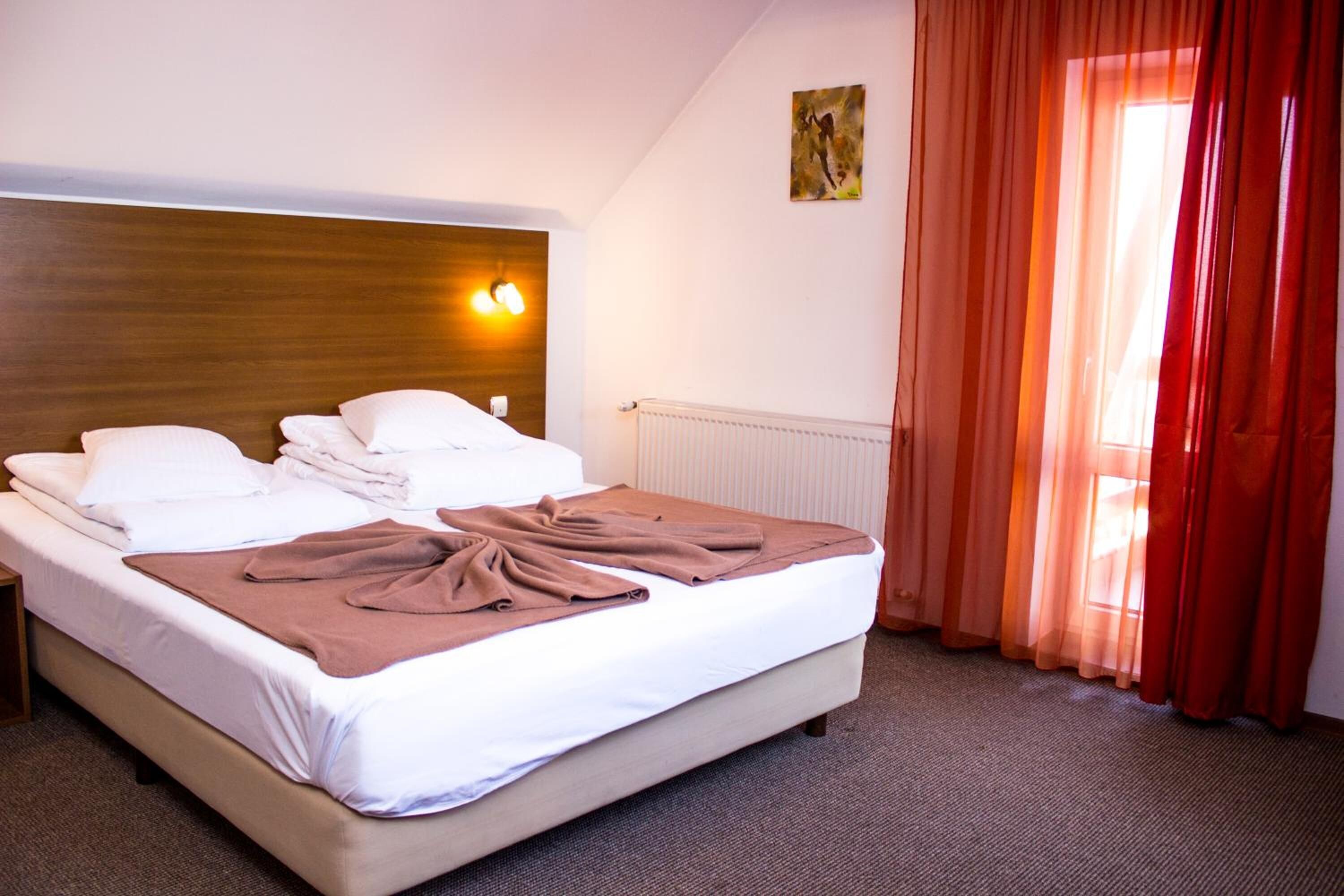 A neatly made bed with white linens and a brown blanket, a wooden headboard, a wall-mounted light, and a window with red curtains.