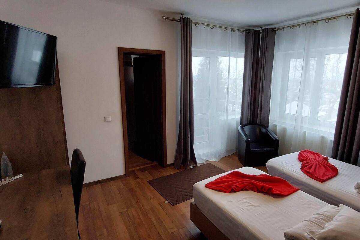 Habitación de hotel con televisión montada en la pared, cama con manta roja, mesita de noche y ventana con cortinas.