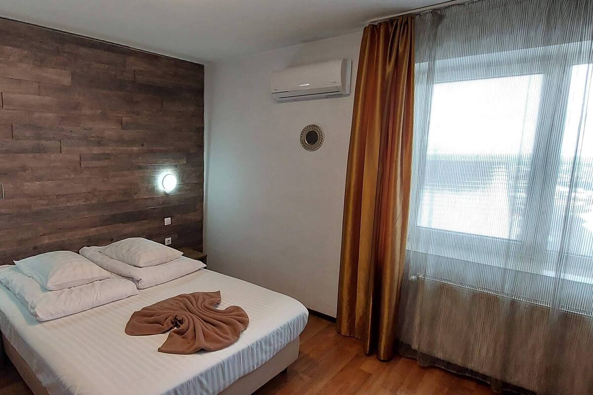 Una cama bien hecha con almohadas y una manta, una pared de madera con iluminación integrada, un equipo de aire acondicionado y una ventana con cortinas.