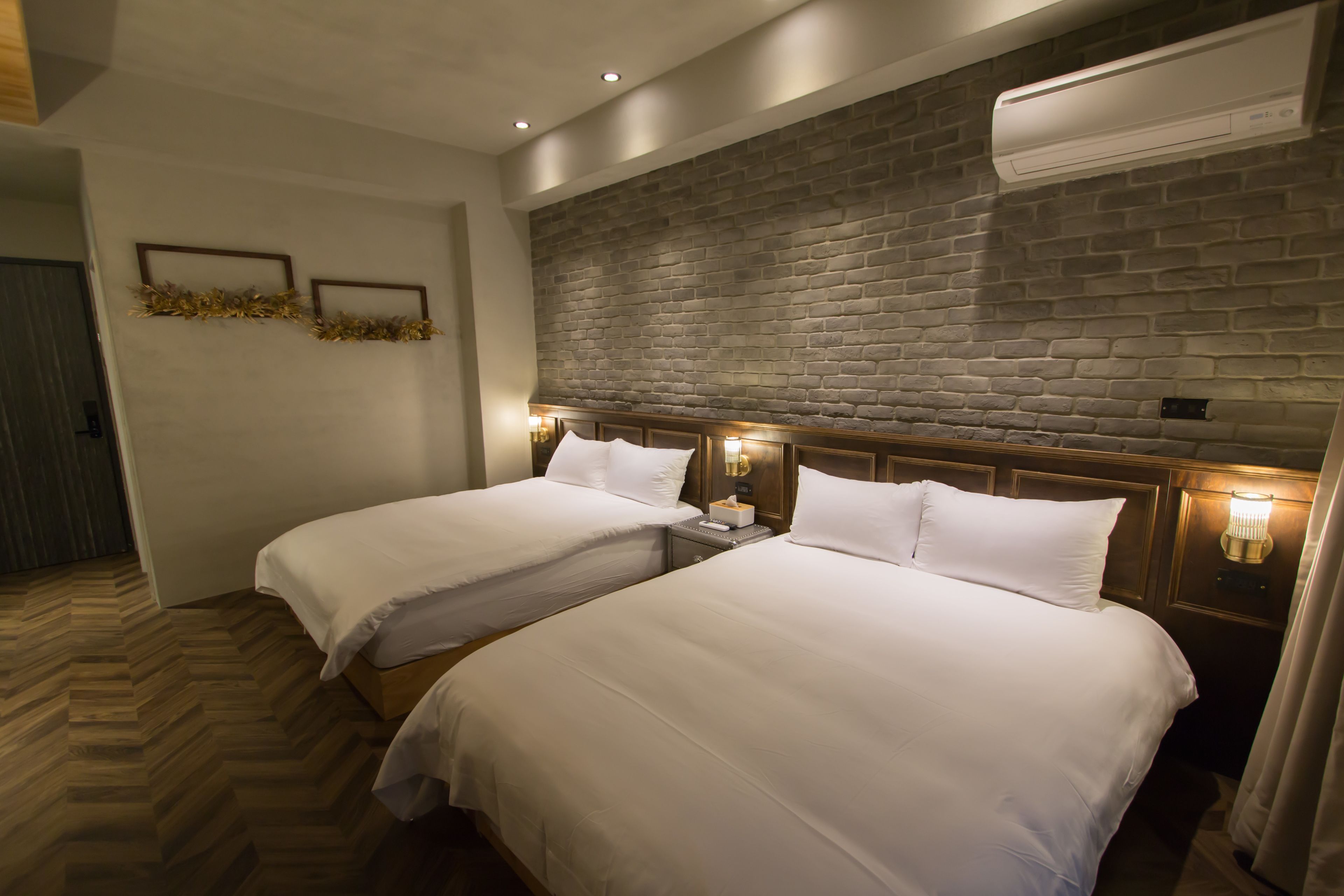 Quarto de hotel com duas camas, piso de madeira, parede de destaque em tijolo e iluminação embutida na parede.