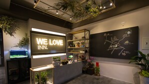 Un área de recepción con un letrero de neón que dice "WE LOVE INN.", un mapa en la pared y un mostrador de recepción con plantas.