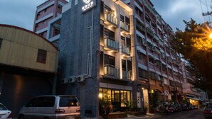 Un hotel de varios pisos con fachada moderna, balcones y un estacionamiento con varios vehículos.