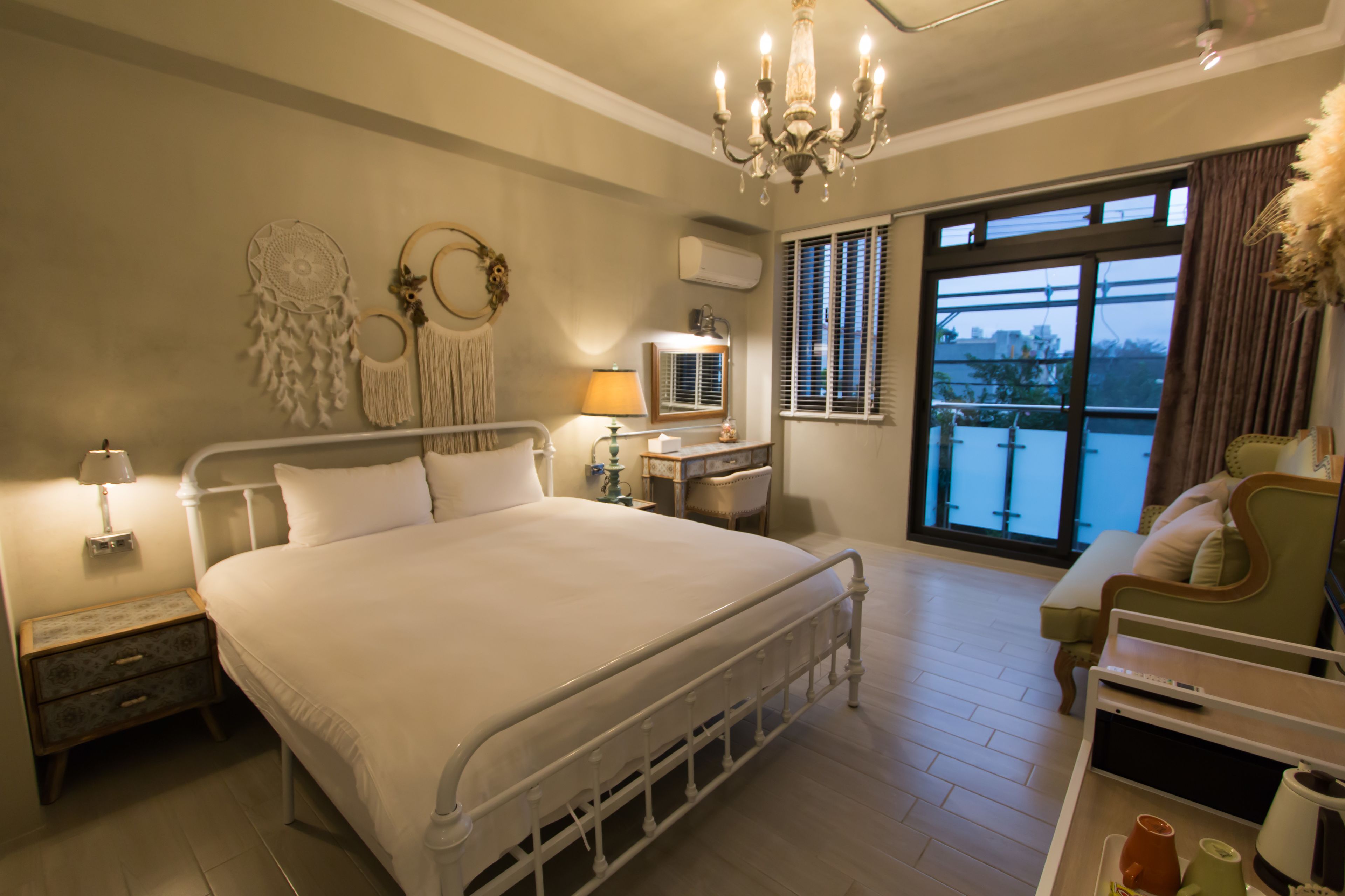 A bedroom with a large bed, bedside tables, a desk, a chair, a chandelier, and a window with blinds.