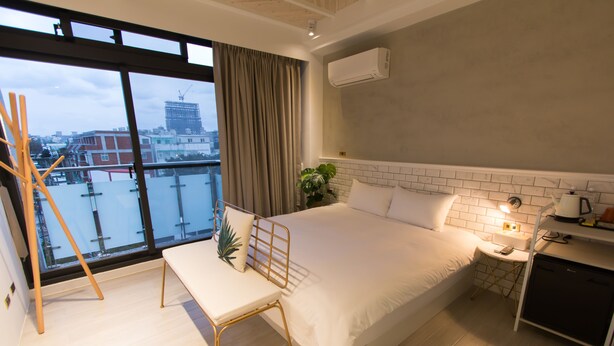 Habitación de hotel moderna con una cama grande, una mesita de noche, un escritorio y vistas a la ciudad a través de amplios ventanales.