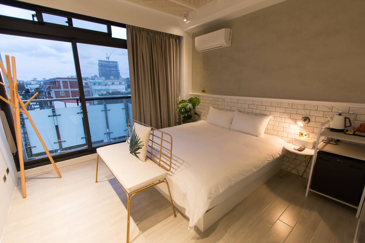 Une chambre d’hôtel moderne dotée d’un grand lit, d’une table de chevet, d’un bureau et offrant une vue sur le paysage urbain grâce à de grandes fenêtres.
