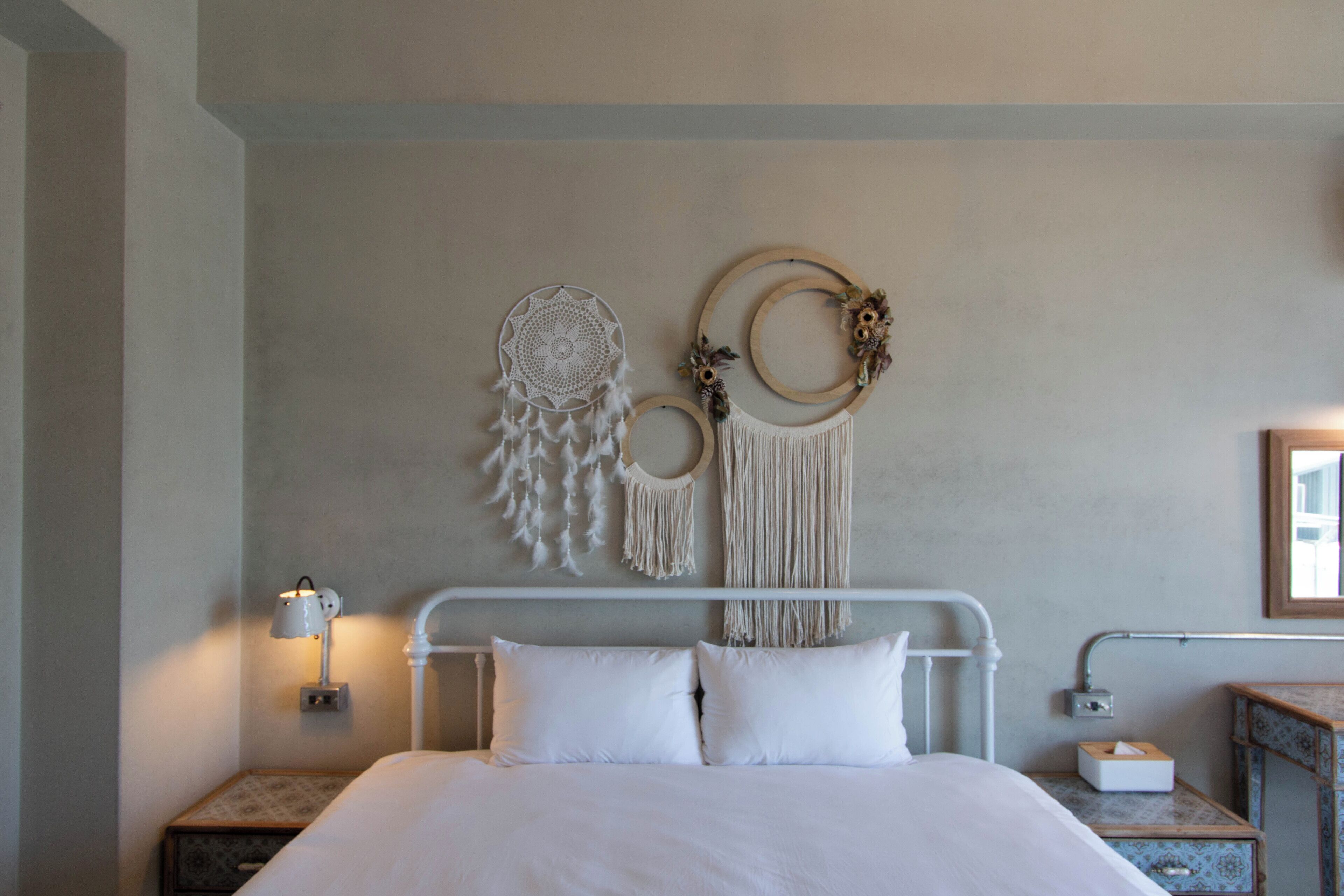 A neatly made bed with white linens, a bedside table with a lamp, and wall decor including a dreamcatcher and macramé wall hanging.