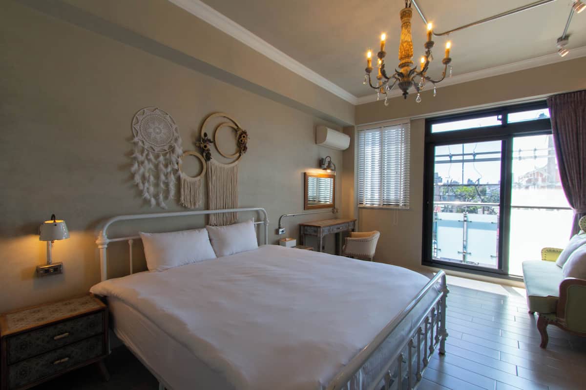 Une chambre spacieuse avec un grand lit, une table de chevet, un bureau et une chaise. On y trouve un lustre, une fenêtre avec des stores et une vue sur l’extérieur.
