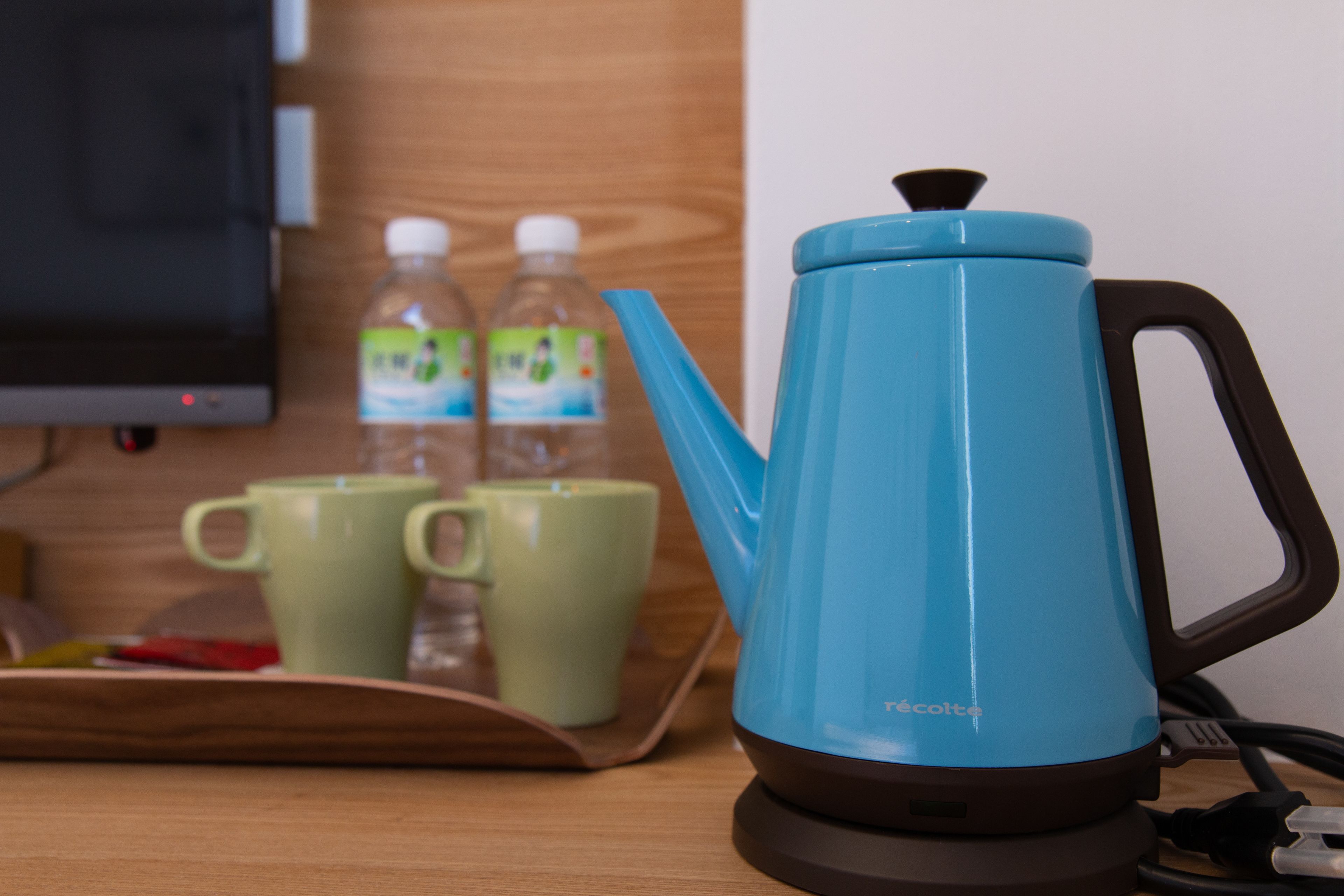 A blue electric kettle on a wooden table with two green cups and two water bottles in the background.