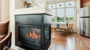 Een moderne woonkamer met een open haard, houten vloeren, een eethoek met een tafel en stoelen, en grote ramen met decoratief glas.