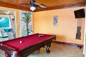 Una mesa de billar en una habitación con techo y paredes de madera, un ventilador de techo, un televisor montado en la pared y un cuadro de palmeras.