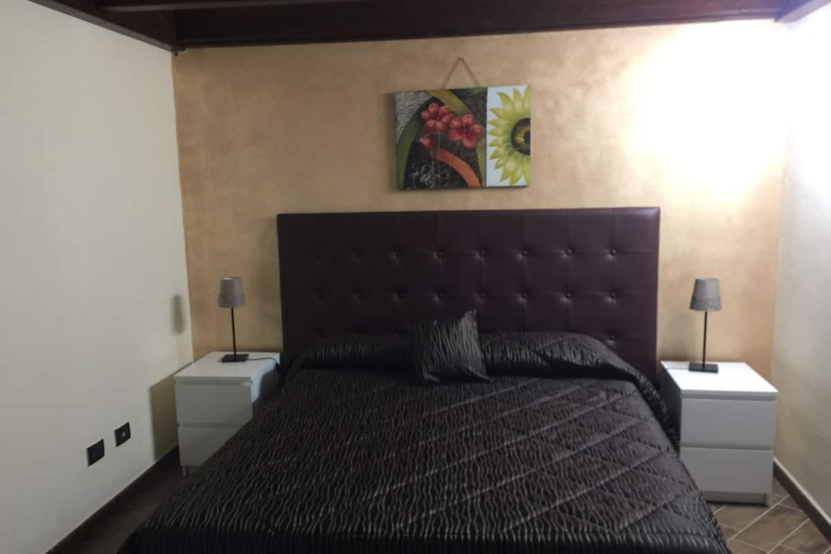 A bedroom with a tufted headboard, a large bed with a patterned bedspread, two bedside tables with lamps, and a wall-mounted painting of flowers.