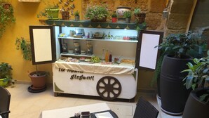 A small outdoor café with a table set for two, a food display case, and various potted plants.