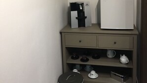 A small kitchenette with a coffee maker, a white refrigerator, a trash bin, and a shelf with cups and a teapot.